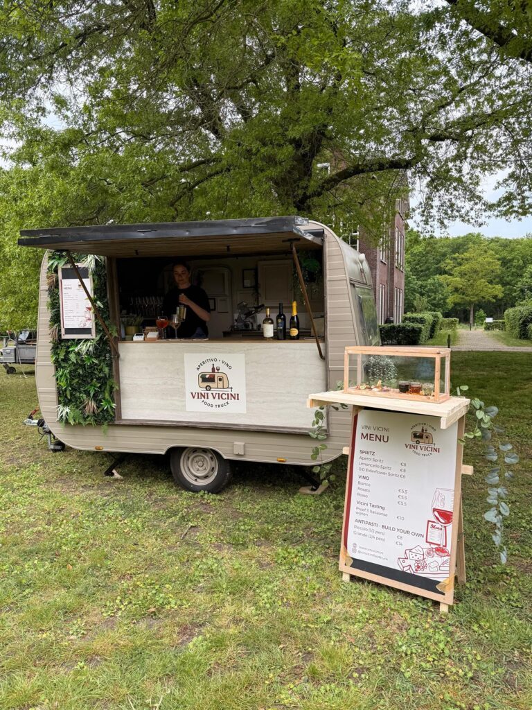 De Vini Vicini caravan met uitgeklapte luifel en menubord onder een grote eik