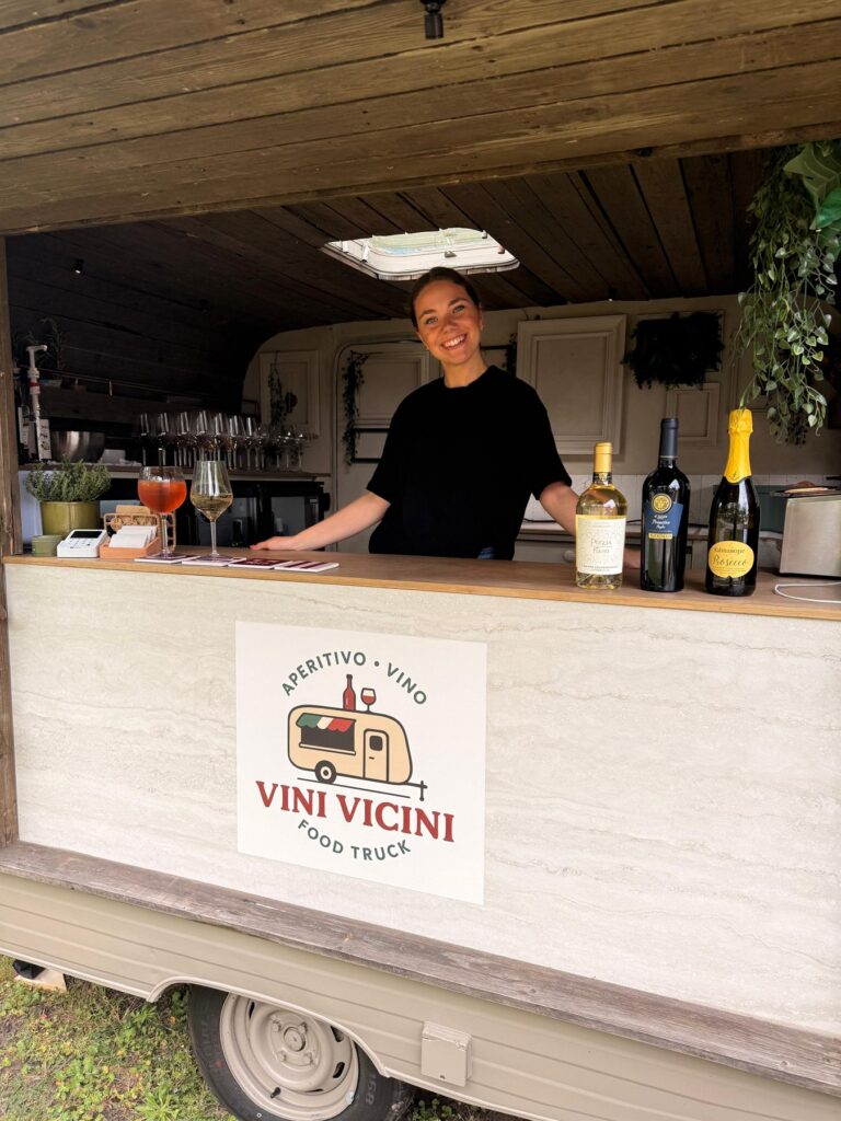 Gastvrouw achter de bar van de Vini Vicini caravan met wijnflessen en spritz
